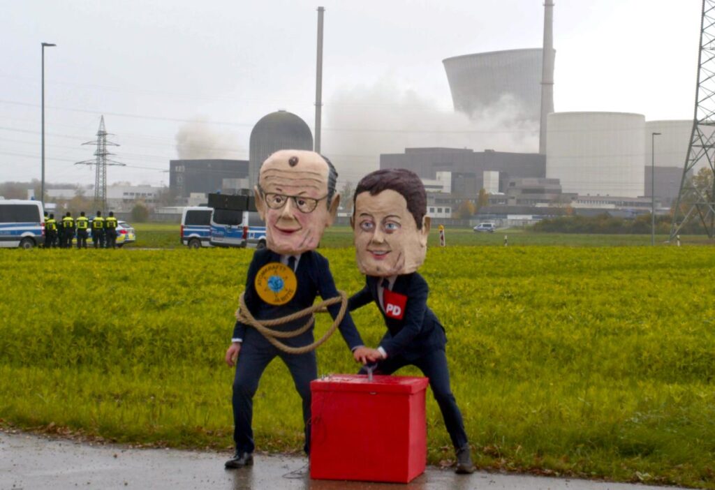Satirische WePlanet-Aktion bei der Sprengung der Kühltürme des Kernkraftwerks Gundremmingen: Aktivisten mit Pappmaché-Köpfen von Friedrich Merz und Lars Klingbeil sprengen die Kühltürme symbolisch.
